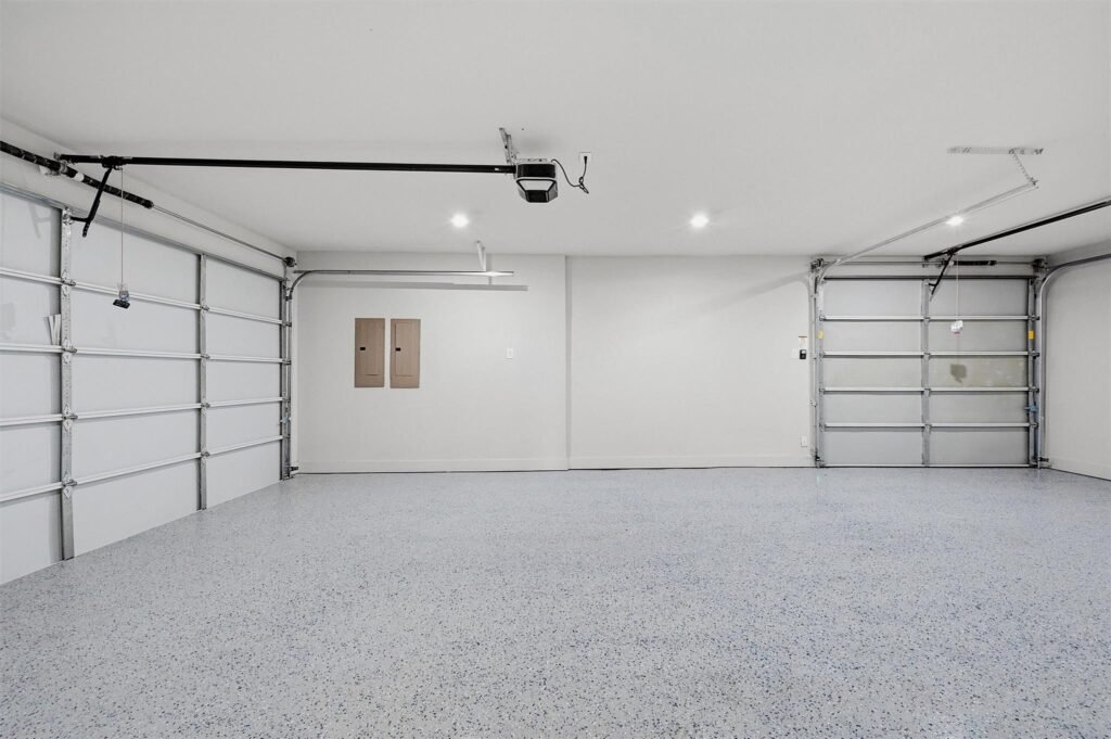 Wide, empty two-car garage with speckled epoxy floor, gray walls, and ceiling-mounted garage door opener along the ceiling track.