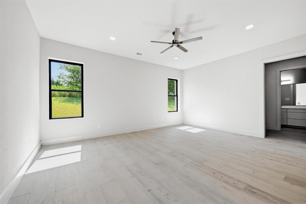 Bright empty living room with light wood floors, white walls, two windows, and a ceiling fan.