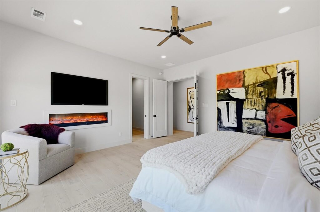 Bright modern bedroom with a ceiling fan, mounted TV, and electric fireplace on a light gray wall.