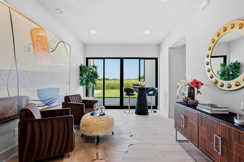 Modern living room with large glass doors open to a green outdoor view, abstract wall art, and a round light-bordered mirror on the right side.