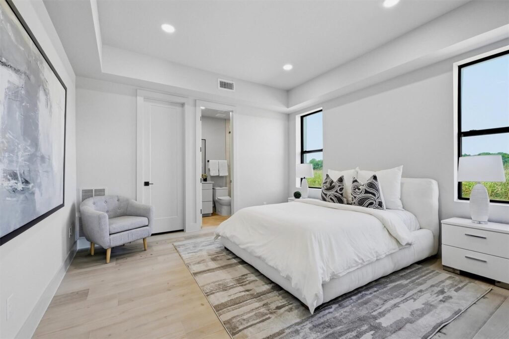 Bright modern bedroom with white bedding, decorative pillows, two nightstands, and large windows; open door to an adjacent bathroom.