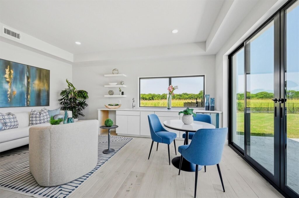 Bright modern living area with a white sofa, blue chairs, and large sliding glass doors opening to a green landscape outside.