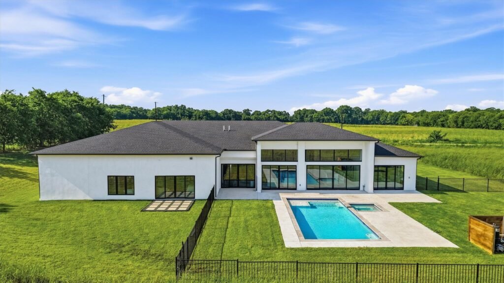 Modern white two-story home with large glass windows, a rectangular pool, and a green lawn in a rural setting.