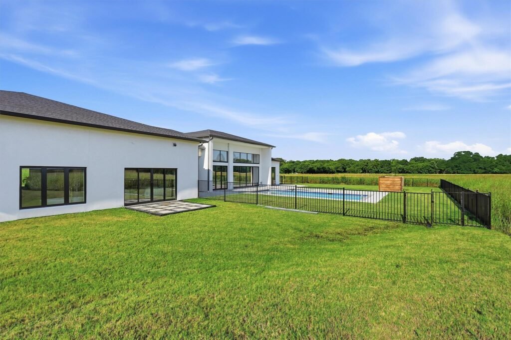 Modern white, single-story home with large glass windows, a fenced backyard, and a rectangular pool on a sunny day.
