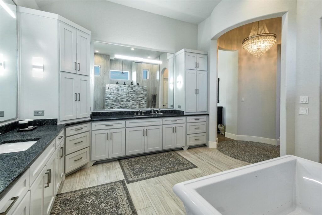 Bright contemporary bathroom with white cabinetry, granite countertops, and a large mirror reflecting a mosaic backsplash and light fixtures in the background.