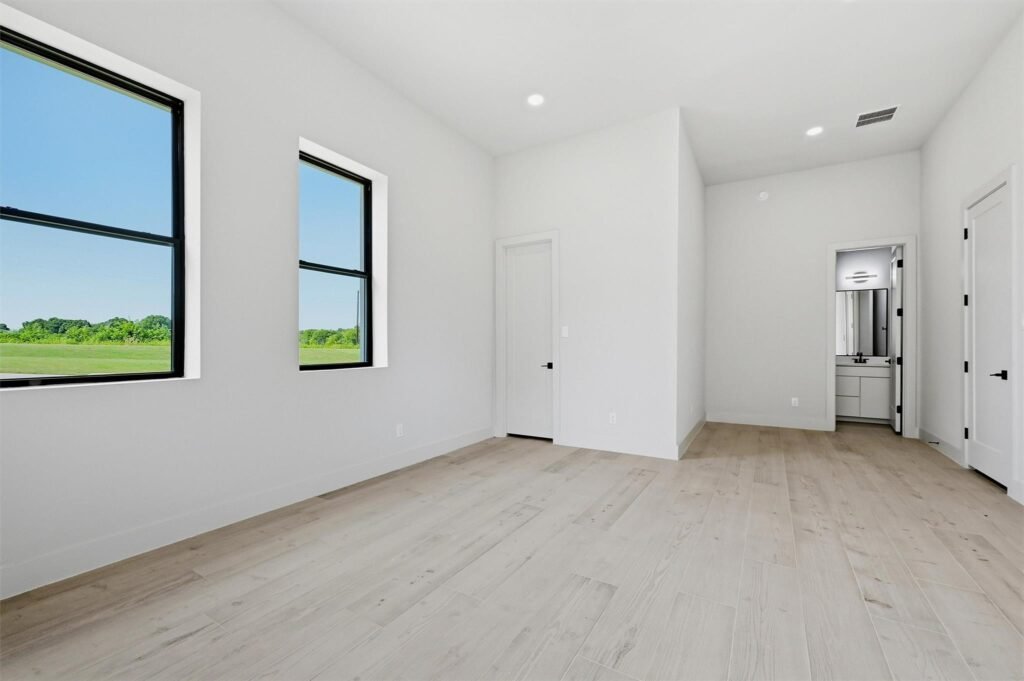 Bright empty room with two large windows showing a green landscape, white walls, and light wood flooring.