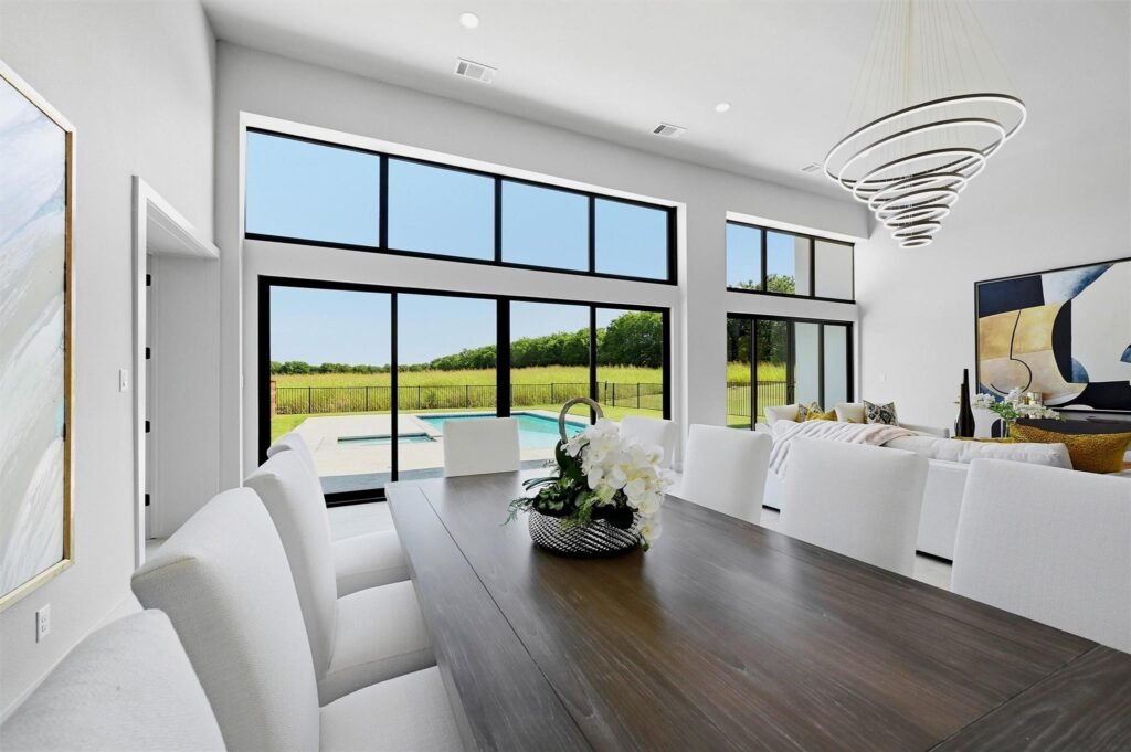 Modern dining room with a long dark wood table, white chairs, and a floral centerpiece overlooking a backyard pool and green lawn.