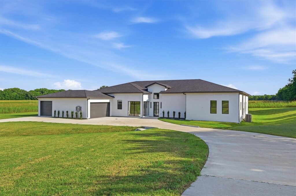 Modern white single-story house with a dark brown roof, large driveway, and green lawn under a blue sky with wispy clouds.