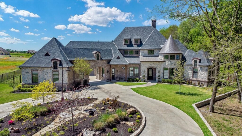 Large stone mansion with slate roofs, circular driveway, and manicured lawn under a blue sky. (informative)