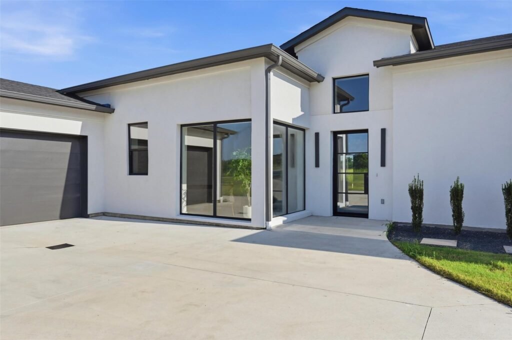 Front view of a modern white house with large glass doors and windows, a gray garage door, and a wide concrete driveway under a blue sky.