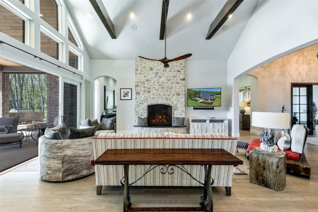 Bright living room with a tall vaulted ceiling, exposed dark wooden beams, a stone fireplace, and a ceiling fan surrounded by comfy seating.