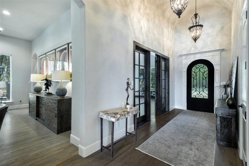 Bright foyer with dark French doors, high ceilings, and a stone-topped console table flanked by two lamps and decorative art pieces by the wall.