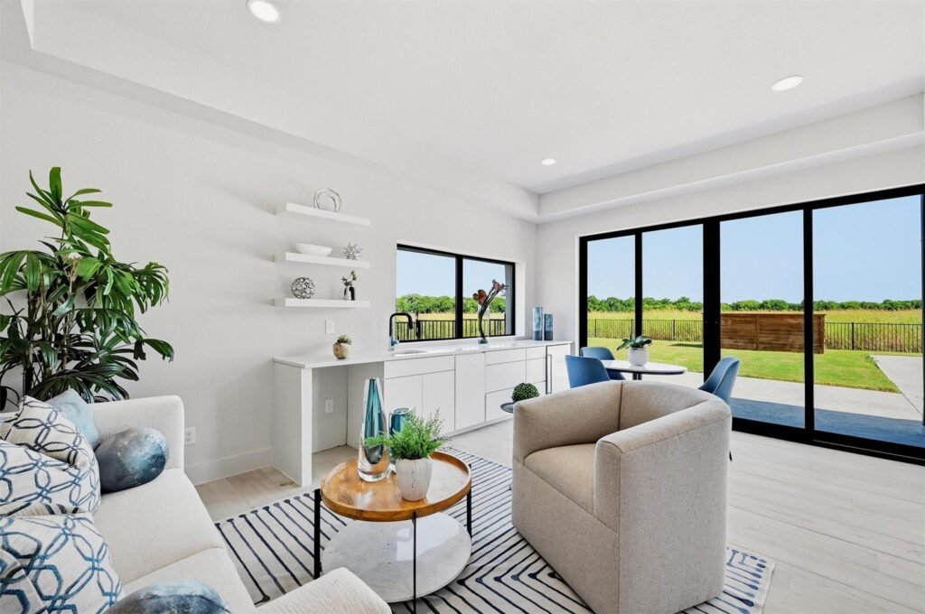 Bright modern living room with large black-framed windows looking out onto a yard; white cabinetry, floating shelves, plants, and blue chairs around round tables.