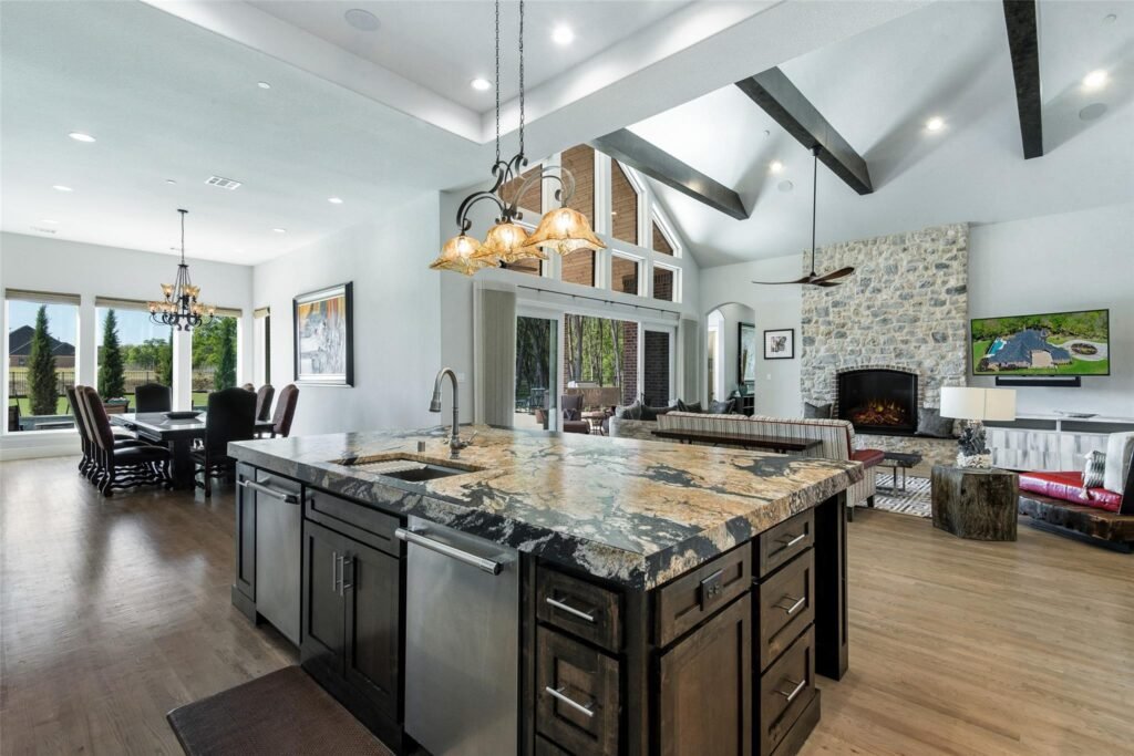 Open-concept kitchen and living room with a dark granite island, pendant lights, and a stone fireplace with a wall-mounted TV nearby.