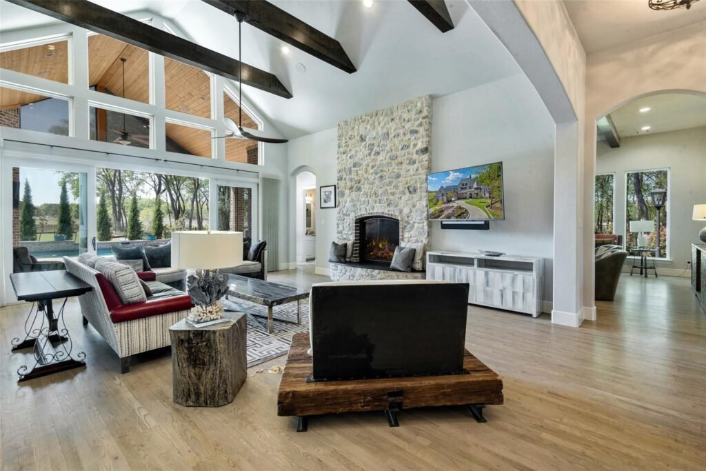Open living room with a tall vaulted ceiling, dark wooden beams, stone fireplace, and a wall-mounted TV by a large window wall.