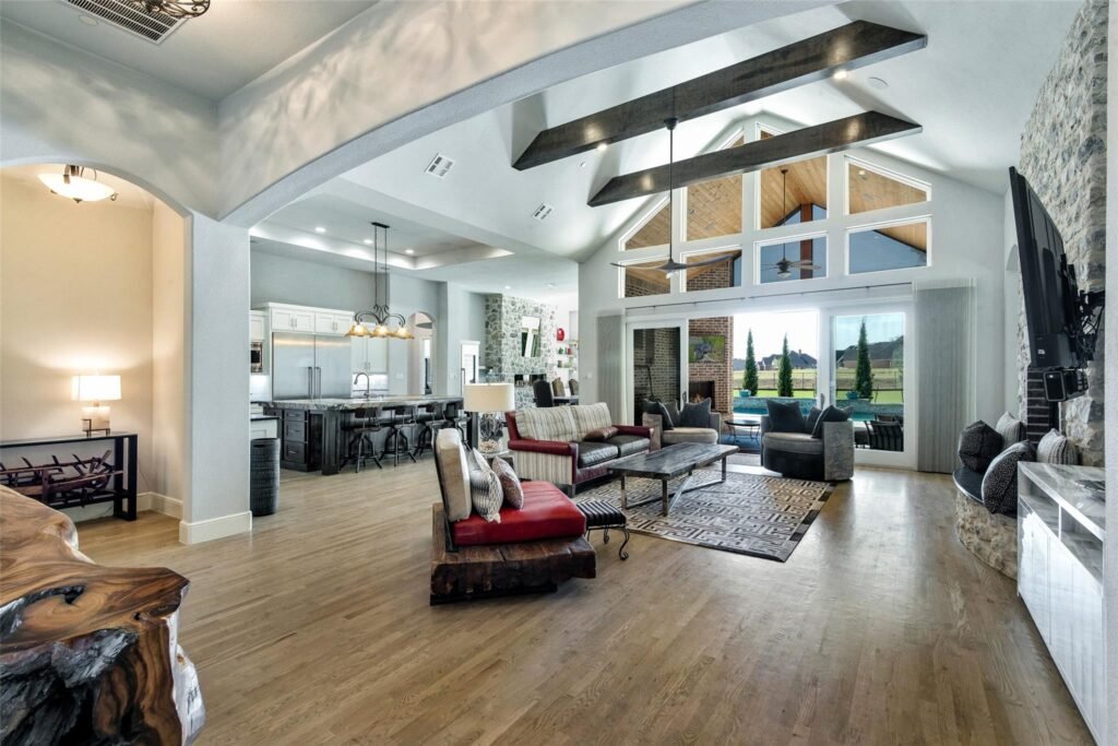 Open living area with vaulted ceiling, exposed dark beams, stone fireplace, and large windows overlooking a backyard yard.