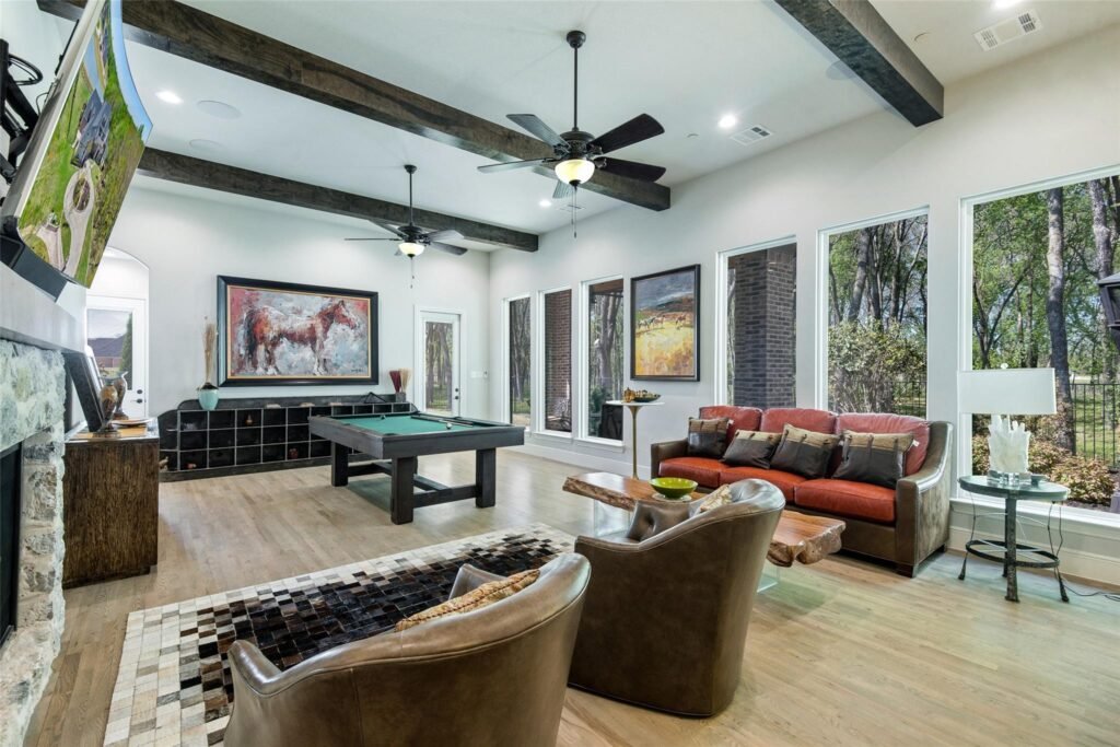 Bright game room with a pool table, leather seating, and large windows revealing greenery outside, plus ceiling fans overhead.