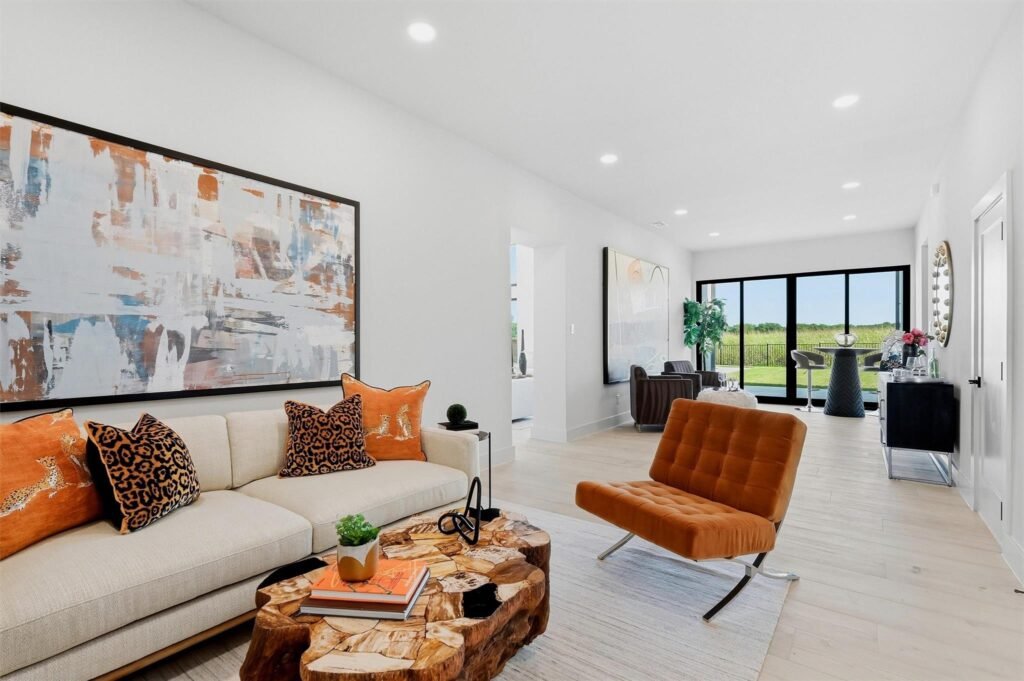 Open living room with a cream sofa, orange chairs, leopard print pillows, and a large abstract painting on the wall; bright natural light from sliding glass doors to a green outdoor view.
