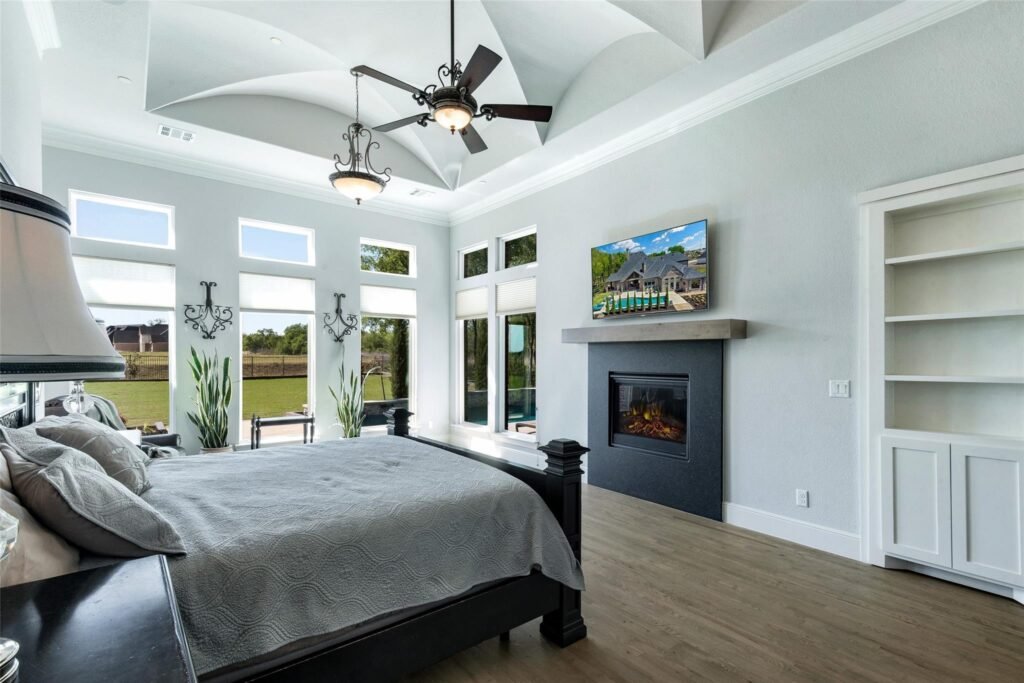 Bright contemporary bedroom with a large bed, vaulted ceiling fans, tall windows, and a wall-mounted TV over a fireplace.