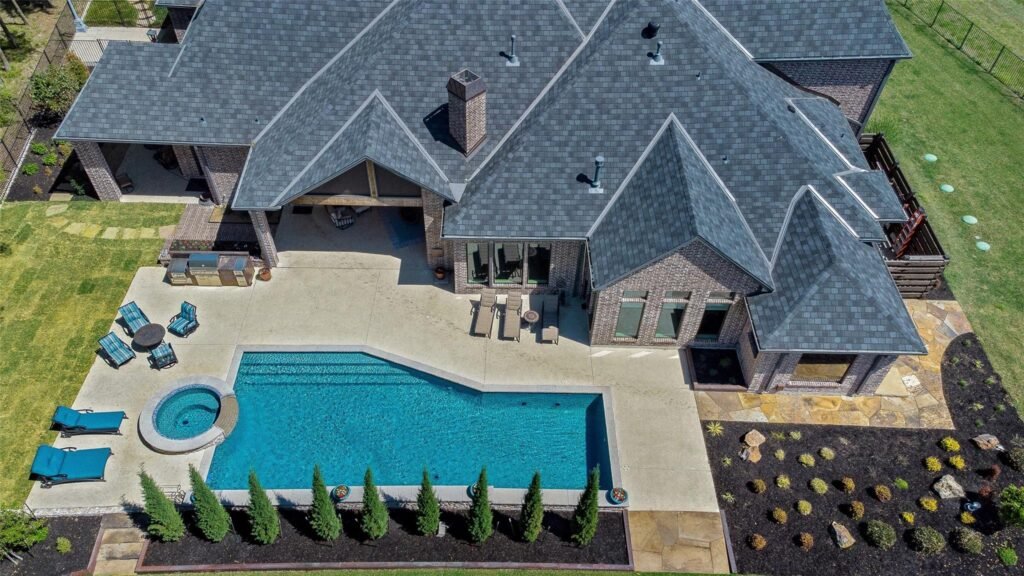 Aerial view of a brick house with gray shingled roof, backyard pool, and outdoor seating area on a concrete patio.