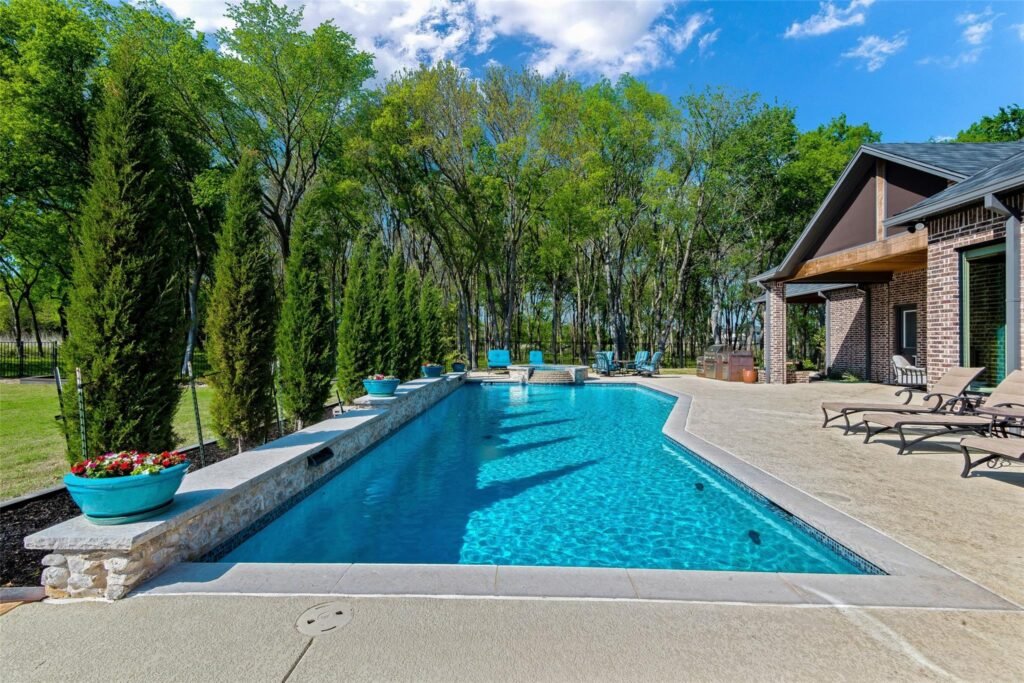 Rectangular backyard pool with stone coping, surrounded by lounge chairs, trees, and a brick house with a covered patio on the right under a blue sky.