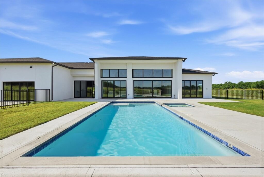 Modern white house with large glass windows and a rectangular pool in the backyard under a blue sky.
