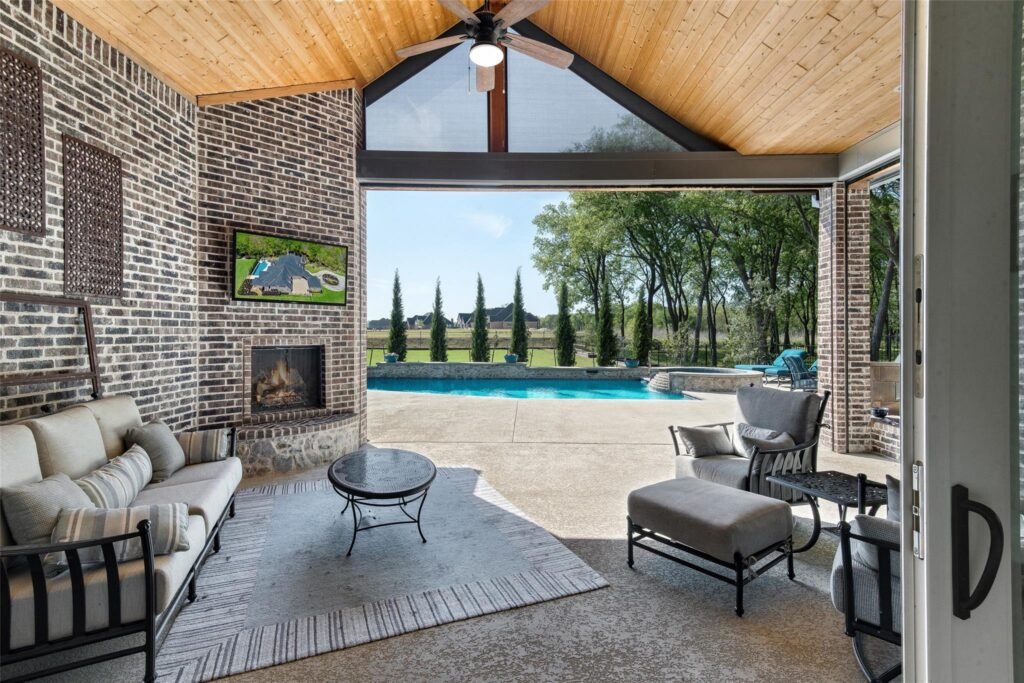 Covered outdoor living area with brick walls, a brick fireplace, and cushioned seating, opening to a backyard pool and green lawn