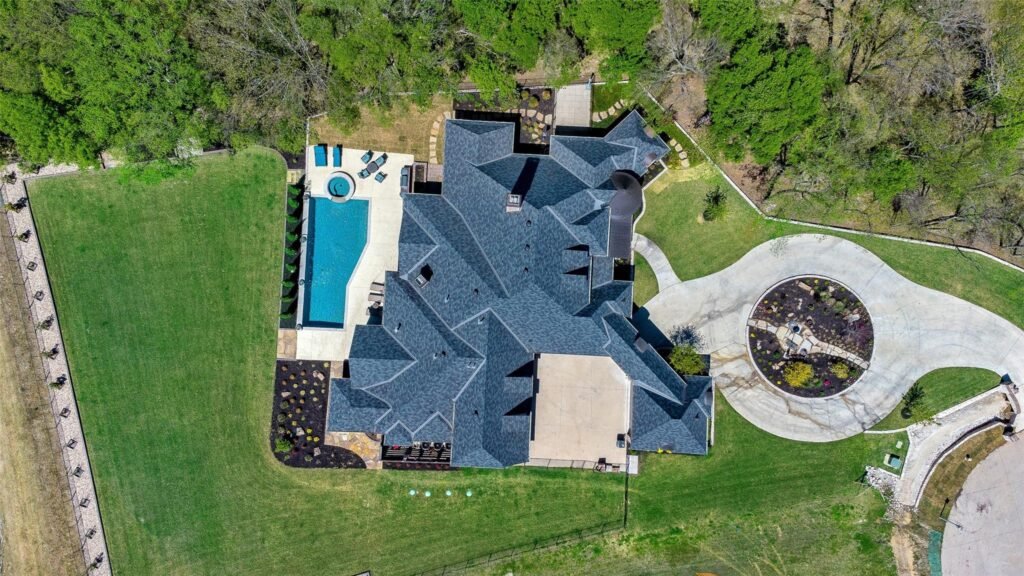 Aerial view of a large gray-roofed house with a rectangular pool, lounge chairs, and a surrounding green lawn and trees.",