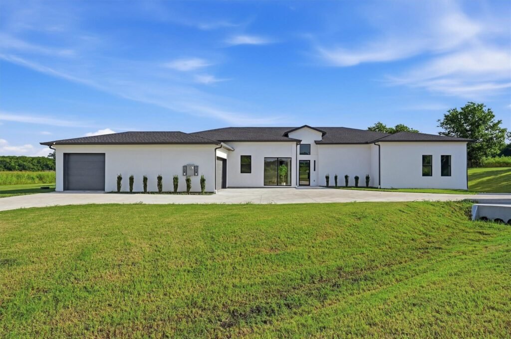 Front view of a modern white house with a dark roof, large windows, and a long driveway on a green lawn under a clear blue sky.