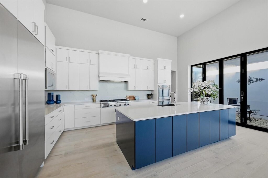 Modern white kitchen with a blue island, stainless appliances, and large glass doors framing the space.