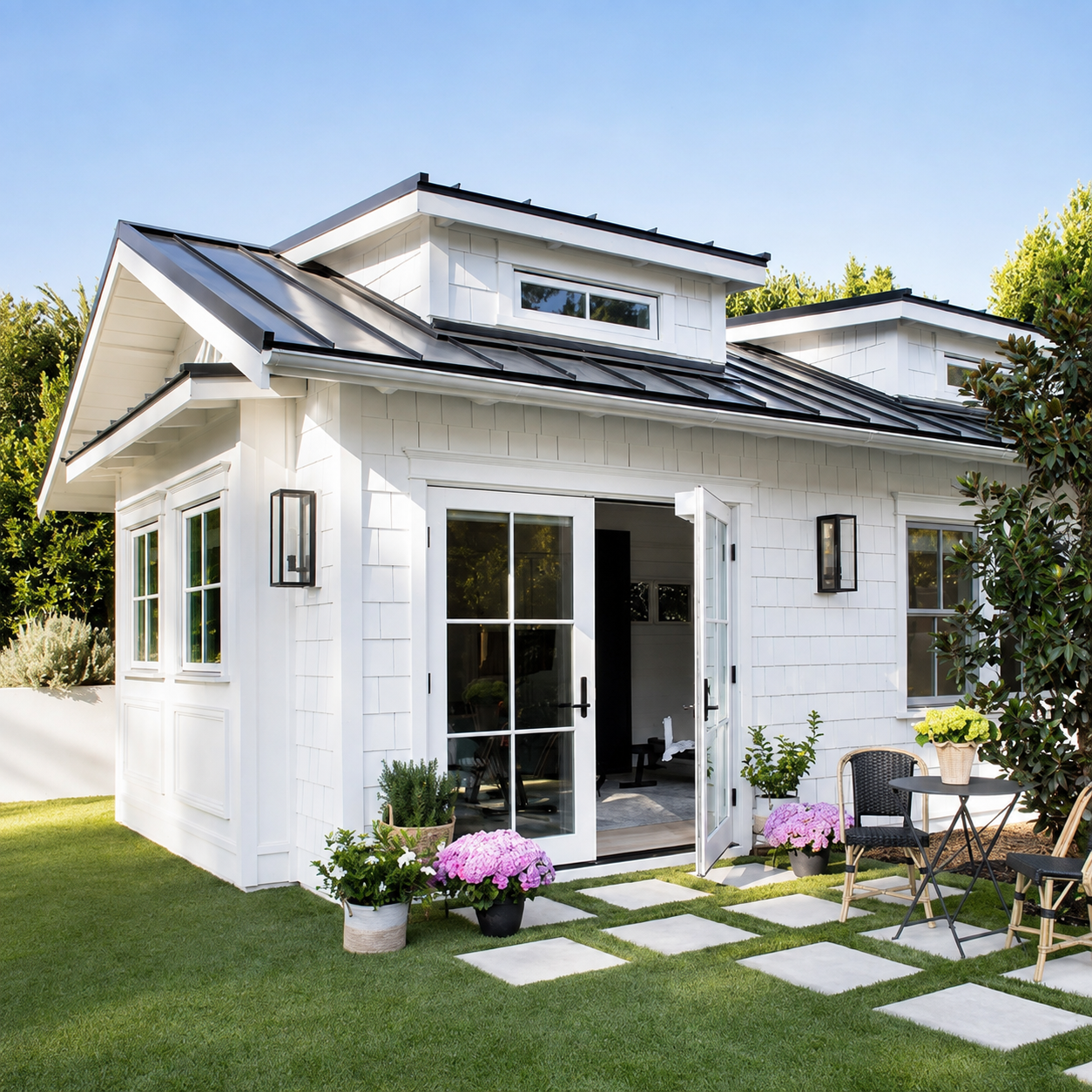 White modern single-story house with open French doors, a green lawn, and a small patio with potted flowers and seating.