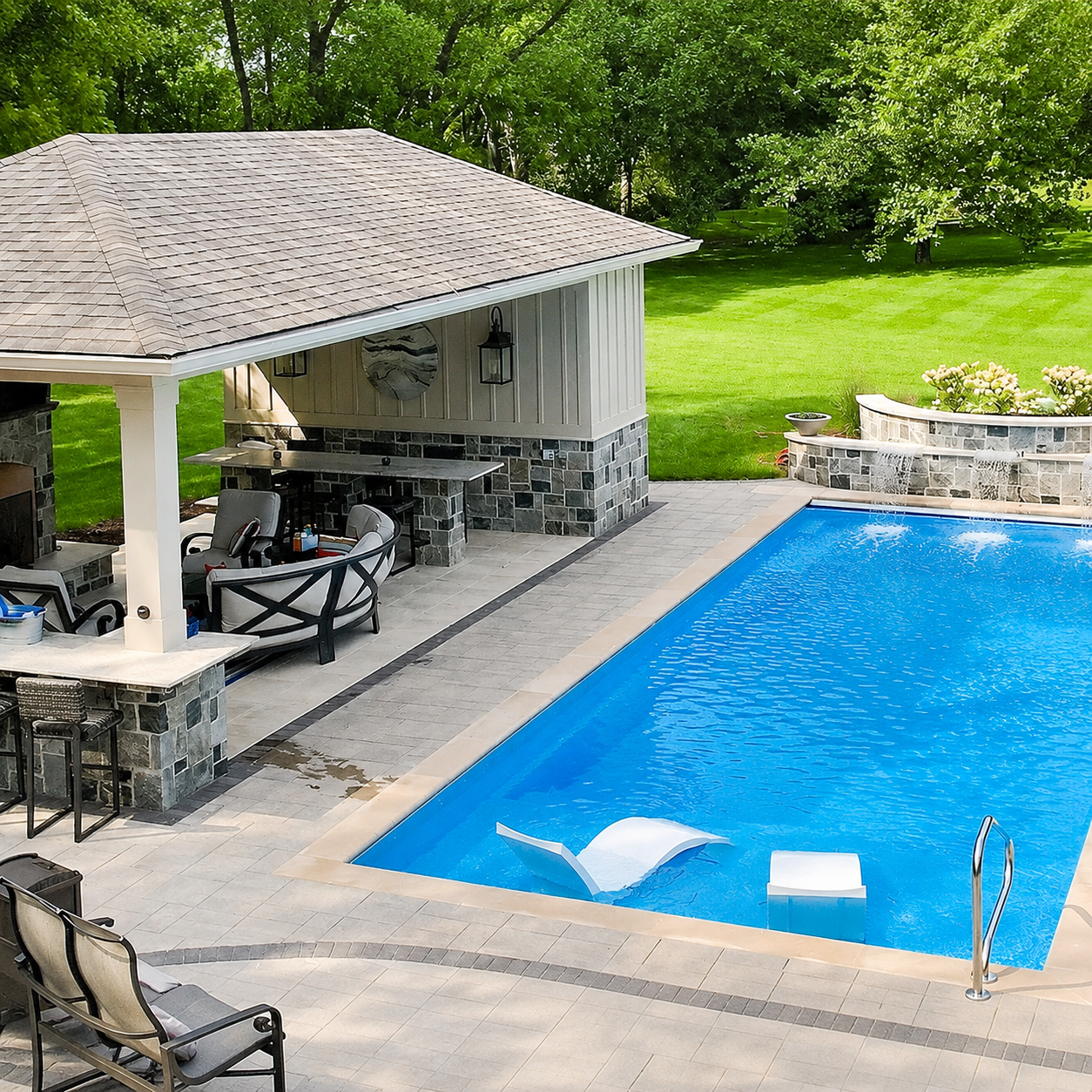 Backyard pool area with a covered stone bar pavilion, lounge chairs, and a bright blue rectangular pool; green lawn in the background.
