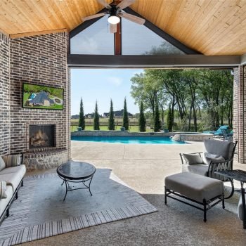 Covered outdoor living area with brick walls, a brick fireplace, and cushioned seating, opening to a backyard pool and green lawn